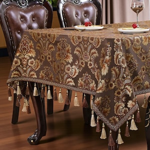 May include: A brown damask tablecloth with a floral pattern, trimmed with tassels. The tablecloth is draped over a wooden table with brown leather chairs. The table is set with plates and wine glasses.