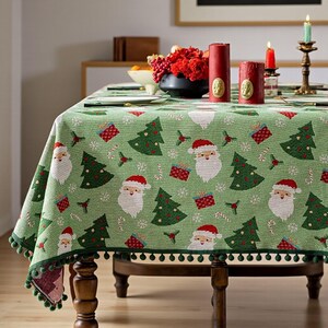 May include: A green Christmas tablecloth featuring Santa Claus, Christmas trees, and gift patterns. The tablecloth has a dark green pom-pom trim. It is set on a wooden table with candles and a floral arrangement, creating a festive atmosphere.