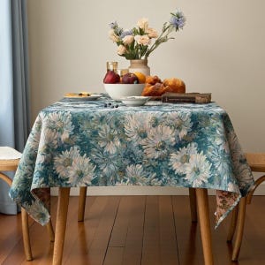 May include: A square tablecloth with a floral pattern in shades of teal, green, and cream. The table is set with a white bowl of fruit, bread, and a vase of flowers. The table is supported by four wooden legs.