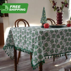 以下が含まれることがあります： 緑の花柄と緑のタッセルが縁取りされた白いテーブルクロスが木製のテーブルを覆っています。小さなライトアップされたクリスマスツリー、赤いリンゴ、赤いベリーが入った花瓶がテーブルにあります。「FREE SHIPPING」という文字が緑色のボックスに入っています。