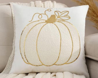 May include: White square decorative pillow with a gold sequined pumpkin design. The pumpkin has a textured appearance and is centered on the pillow. The pillow is on a cream-colored sofa with a white knitted throw.