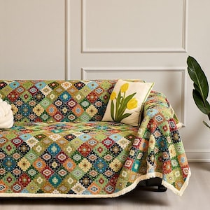 Puede incluir: Un sofá cubierto con una manta estampada colorida. La manta presenta un diseño geométrico en tonos verdes, rojos, azules y amarillos. Un cojín decorativo con bordado de flores amarillas descansa sobre el sofá.