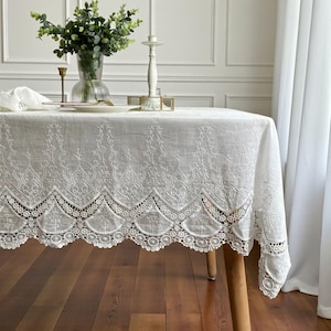 May include: A white rectangular tablecloth with intricate embroidered floral patterns and a scalloped lace edge. The table is set with a vase of greenery, candles, and a plate. The tablecloth is draped over a wooden table.