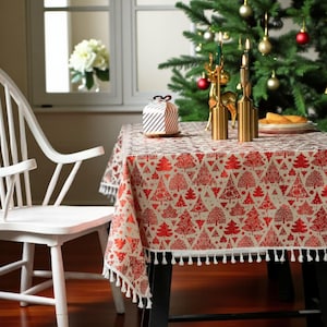 May include: A festive table setting featuring a tablecloth with a red Christmas tree pattern. Gold candle holders and a decorative gift box adorn the table. A white chair and Christmas tree are in the background.