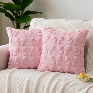Puede incluir: Dos cojines decorativos rosas con un patrón de lazo. Los cojines están hechos de un material suave y texturizado y están colocados en un sofá color crema. Los cojines añaden un toque de elegancia a cualquier espacio habitable.