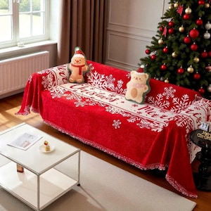 May include: A red sofa cover with a white snowflake design and the word "Christmas". Two decorative cushions, one a gingerbread man and the other a bear, are on the sofa. A decorated Christmas tree is visible in the background.
