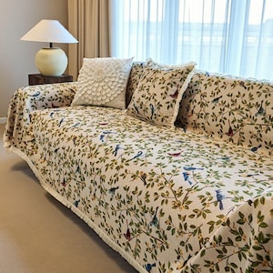 以下が含まれることがあります： クリーム色のソファカバーには、緑の葉とカラフルな鳥の模様が施されています。スローにはフリンジエッジが付いています。花柄と鳥柄の装飾枕がルックを完成させます。白いランプとサイドテーブルが背景に見えます。