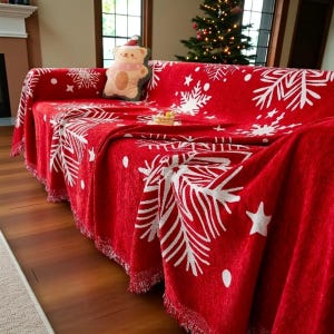 Könnte beinhalten: Eine rote Sofadecke mit einem weißen Schneeflocken- und Sternmuster. Die Decke hat einen Fransenrand und ist über ein Sofa drapiert. Ein Teddybär-Kissen mit Weihnachtsmütze liegt auf dem Sofa.