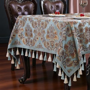May include: A light blue damask tablecloth with a floral pattern in shades of brown and gold. The tablecloth has a decorative border and is trimmed with tassels in light blue and cream colours. The tablecloth is draped over a dark wooden table.