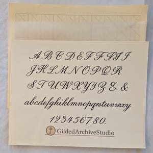 May include: A cream-colored card displays a handwritten alphabet in elegant cursive script, including uppercase and lowercase letters, numbers, and the ampersand symbol. The card also features the text "GildedArchiveStudio" and a logo.