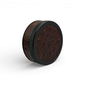 May include: A round, brown leather grinder with a black metal rim. The leather has a textured, ostrich-like pattern. The grinder is designed with a ribbed edge for grip and a smooth, flat top.