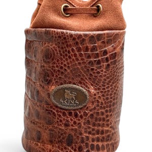 May include: A brown leather drawstring bag with a crocodile texture on the lower portion and a suede top. The bag features a metal emblem with a bull and the text "SKINS GENUINE SOVIES". The bag is cylindrical in shape.