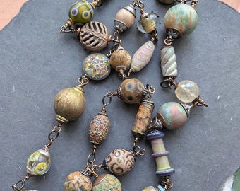 May include: A handcrafted necklace featuring a variety of artisan beads in earthy tones. The beads include round, oval, and cylindrical shapes with intricate patterns and textures, strung together with copper wire.
