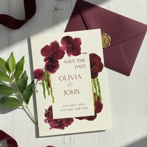 Puede incluir: Una tarjeta crema de "Save the Date" con detalles florales burdeos y texto. La tarjeta presenta los nombres "Olivia & John" y la fecha "1 de enero de 2028, Nueva York, Nueva York". También hay un sobre burdeos con un sello de cera dorado.
