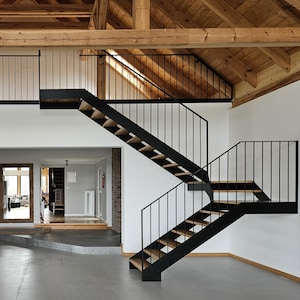 Może przedstawiać: Widok wnętrza nowoczesnego domu z antresolą i schodami. Przestrzeń charakteryzuje się szarą betonową podłogą, białymi ścianami i odsłoniętymi drewnianymi belkami. Czarne metalowe schody prowadzą na antresolę. Widoczny jest stół jadalny i krzesła.