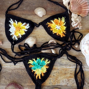 May include: Black crochet bikini set with floral accents in yellow, orange, and white. The bikini top and bottom feature a flower design. The set is displayed on a wooden surface with seashells.