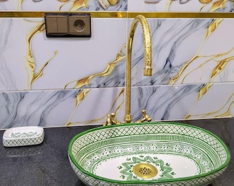 May include: An oval ceramic sink with a green and white geometric pattern. The sink is set on a black countertop with a gold faucet. A matching soap dish is to the left. The background features decorative tiles.