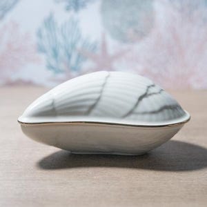 May include: A white ceramic seashell-shaped trinket box with a gold trim. The lid has a textured surface with grey marbling. The box is closed and sits on a light-coloured surface.