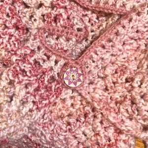 May include: Close-up of a pink and white crocheted fabric with a decorative button featuring a pink and white floral design.
