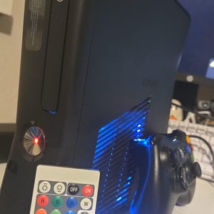 Könnte beinhalten: Eine schwarze Xbox 360 Spielkonsole mit leuchtend rotem Power-Knopf und blauer Innenbeleuchtung, begleitet von einem schwarzen kabellosen Gamecontroller und einer weißen RF-Fernbedienung mit verschiedenen Farb- und Modustasten.