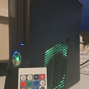Könnte beinhalten: Eine schwarze Xbox 360 Spielkonsole mit einem leuchtend grünen Einschaltknopf und grüner Innenbeleuchtung, die durch die seitlichen Lüftungsschlitze sichtbar ist, sowie eine weiße, rechteckige RF-Fernbedienung mit bunten Tasten.