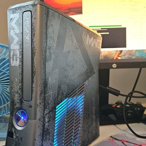 Könnte beinhalten: Eine limitierte graue Xbox 360 Konsole mit abgenutzter Metalloptik und Modern Warfare 3 Branding. Die Frontplatte zeigt das MW3-Logo und den Xbox 360-Schriftzug, wobei der kreisförmige Einschaltknopf von einem hellblauen Licht beleuchtet wird.