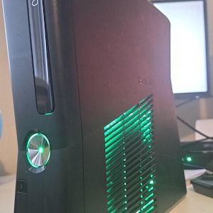 Könnte beinhalten: Eine schwarze Xbox 360 Spielkonsole in vertikaler Ausrichtung. Die Vorderseite zeigt das Xbox 360 Logo und einen kreisförmigen Einschaltknopf, der hellgrün leuchtet. Ein seitliches Lüftungsgitter gibt den Blick auf ein leuchtend grünes Licht frei.