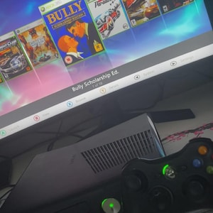 Könnte beinhalten: Eine Xbox 360-Konsole mit einem schwarzen Controller und einem Bildschirm, der mehrere Spielcover anzeigt, darunter "Bully Scholarship Ed." und "Gears of War." Die Konsole ist schwarz und silber, mit einer grünen Power-Leuchte.