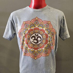 May include: A gray t-shirt featuring a colorful mandala design with the Om symbol at the center. The mandala is in shades of red, yellow, and brown. The shirt is displayed on a mannequin.