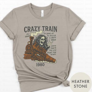 May include: Heather stone-colored t-shirt with a graphic of a train and a skeleton conductor. The text reads "CRAZY TRAIN" and "1980". Additional text includes "I'm going off the rails on a crazy train" and "Learn how to love and forget how to hate."