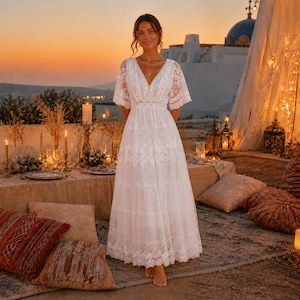 May include: A floor-length white dress featuring short lace sleeves, a deep V-neckline, and a tiered skirt with intricate floral lace detailing throughout the bodice and hem. The garment has a fitted waist and a flowing, layered silhouette.