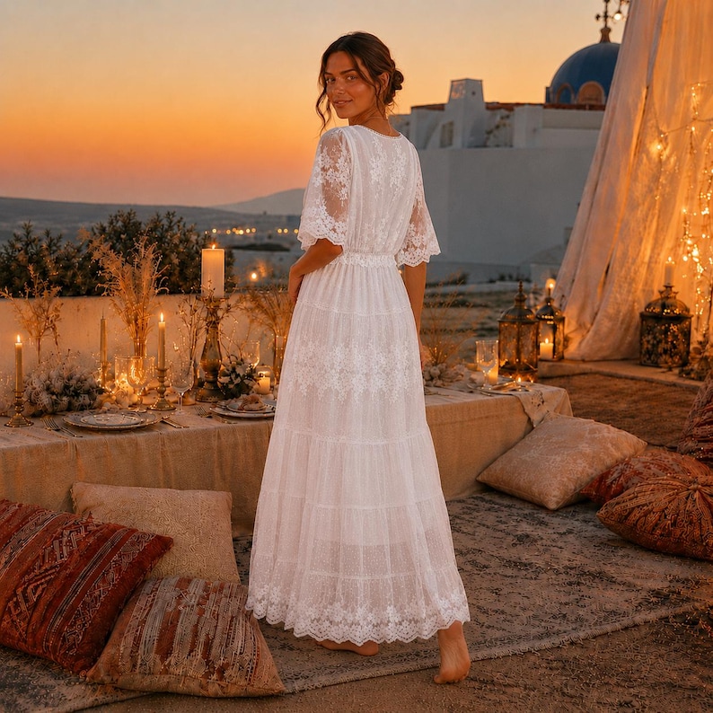 May include: A floor-length white lace dress featuring short sleeves, a cinched waist, and a tiered skirt with intricate floral embroidery patterns throughout the fabric. The garment has a V-neckline and a sheer lace overlay design.