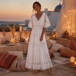 May include: A floor-length white dress featuring a V-neckline, short flutter sleeves, and a tiered skirt. The garment is crafted from sheer lace fabric with intricate floral embroidery patterns throughout the bodice, sleeves, and hemline.