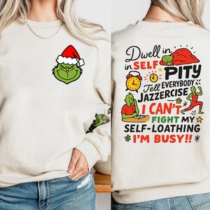 May include: A cream-colored sweatshirt featuring a Grinch face wearing a Santa hat on the front. The back of the sweatshirt has the text "Dwell in Self Pity" and other phrases, along with Grinch-themed illustrations. The sweatshirt is a festive holiday item.