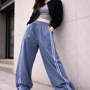 May include: A woman's outfit featuring a black jacket, gray crop top, and blue sweatpants with white stripes. The outfit is completed with white sneakers. The pants have a logo on the left leg. The woman is leaning against a gray wall.