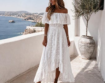 May include: A white, off-the-shoulder dress with intricate eyelet embroidery. The dress features a ruffled neckline and a high-low hem. The dress is paired with beige sandals. The background shows a coastal scene with blue sea and a clear sky.