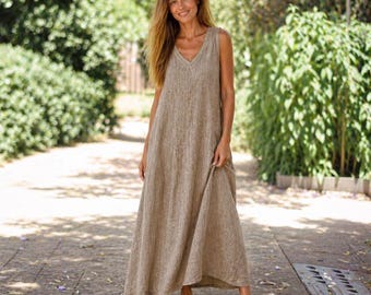 May include: A beige, sleeveless maxi dress with a V-neck. The dress has a loose fit and falls to the floor. The person is wearing brown sandals. The background features green foliage and a paved path.