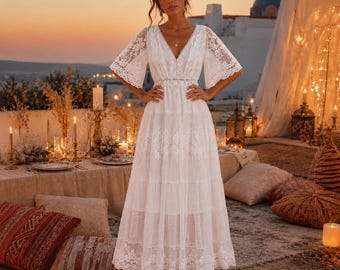 May include: A floor-length white dress featuring intricate lace detailing, a deep V-neckline, and wide, flowing sleeves. The garment has a tiered skirt design with delicate floral embroidery patterns throughout the fabric and a cinched waistline.