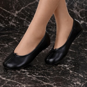 Peut inclure: Ballerines noires avec un bout arrondi et un petit nœud décoratif. Les chaussures sont faites d'une matière lisse et ont un petit talon. Les ballerines sont présentées sur une surface en marbre.