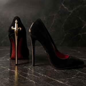 May include: Two black high heels with gold detailing. The shoes have pointed toes and stiletto heels. The soles are red. One heel features a gold sword-like design, the other a gold zip. The heels are on a dark surface.