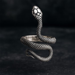 Peut inclure: Bague argentée en forme de serpent, enroulée autour du doigt. La tête du serpent est relevée, avec un motif d'écailles détaillé. La bague a une finition texturée et vieillie, créant un bijou saisissant.