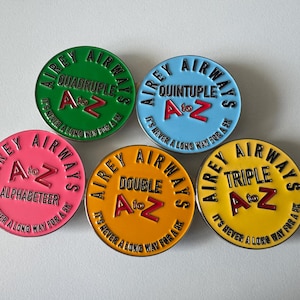 Op de afbeelding: Vijf kleurrijke Airey Airways emaille pins. Elke pin is rond met een andere kleur achtergrond: groen, blauw, roze, oranje en geel. Elke pin heeft de tekst "Airey Airways" en "A to Z" met een ander woord zoals "Quadruple" of "Alphabeteer".