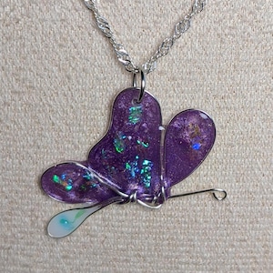 May include: A purple butterfly pendant necklace with a silver chain. The butterfly has iridescent glitter and a silver wire outline. The pendant's lower portion has a white and blue teardrop shape.