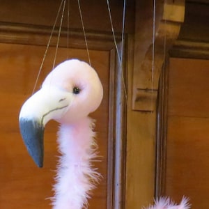 Könnte beinhalten: Eine rosa Flamingo-Puppe mit weißem Schnabel und Beinen. Der Flamingo besteht aus weichen, flauschigen Federn und wird von dünnen Fäden gehalten. Der Hintergrund ist eine Holztür.