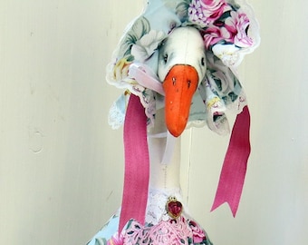 Puede incluir: Una caprichosa figura de ganso vestida con un vestido y un gorro con estampado floral. El ganso tiene pico y patas naranjas y lleva una pequeña cesta de mimbre. El vestido y el gorro presentan un fondo azul claro con flores rosas y moradas.