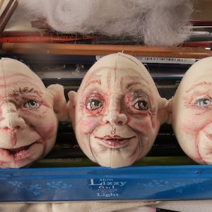 Puede incluir: Tres cabezas de muñeca de tela sin terminar con rasgos pintados. Las cabezas son blancas con mejillas rosadas y tienen ojos, narices y bocas pintados. Las cabezas están sentadas en un recipiente de metal azul con las palabras "Lizzy Cin 1 Light" impresas en el lateral.