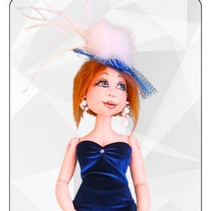 Könnte beinhalten: Ein blaues Samtkleid mit einem trägerlosen Ausschnitt und einer weißen Clutch. Die Puppe trägt einen weißen Hut mit blauen Verzierungen und hat rote Haare. Der Text "Krista by Sharon Mitchell" befindet sich unter der Puppe.