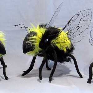 May include: Three plush bumblebees with black and yellow fur, clear wings, and black legs. The bumblebees are arranged in a row on a white surface.