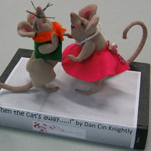 May include: Two gray felt mouse figurines dancing on a book titled "When the Cat's Away..." by Dan Cin Knightly. The female mouse wears a pink dress with a pearl necklace and the male mouse wears a green vest with an orange scarf.