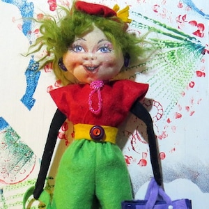 Puede incluir: Una muñeca hecha a mano con cabello verde, un atuendo rojo y amarillo y un bolso morado. La muñeca tiene una cara pintada con mejillas sonrosadas y una gran sonrisa.